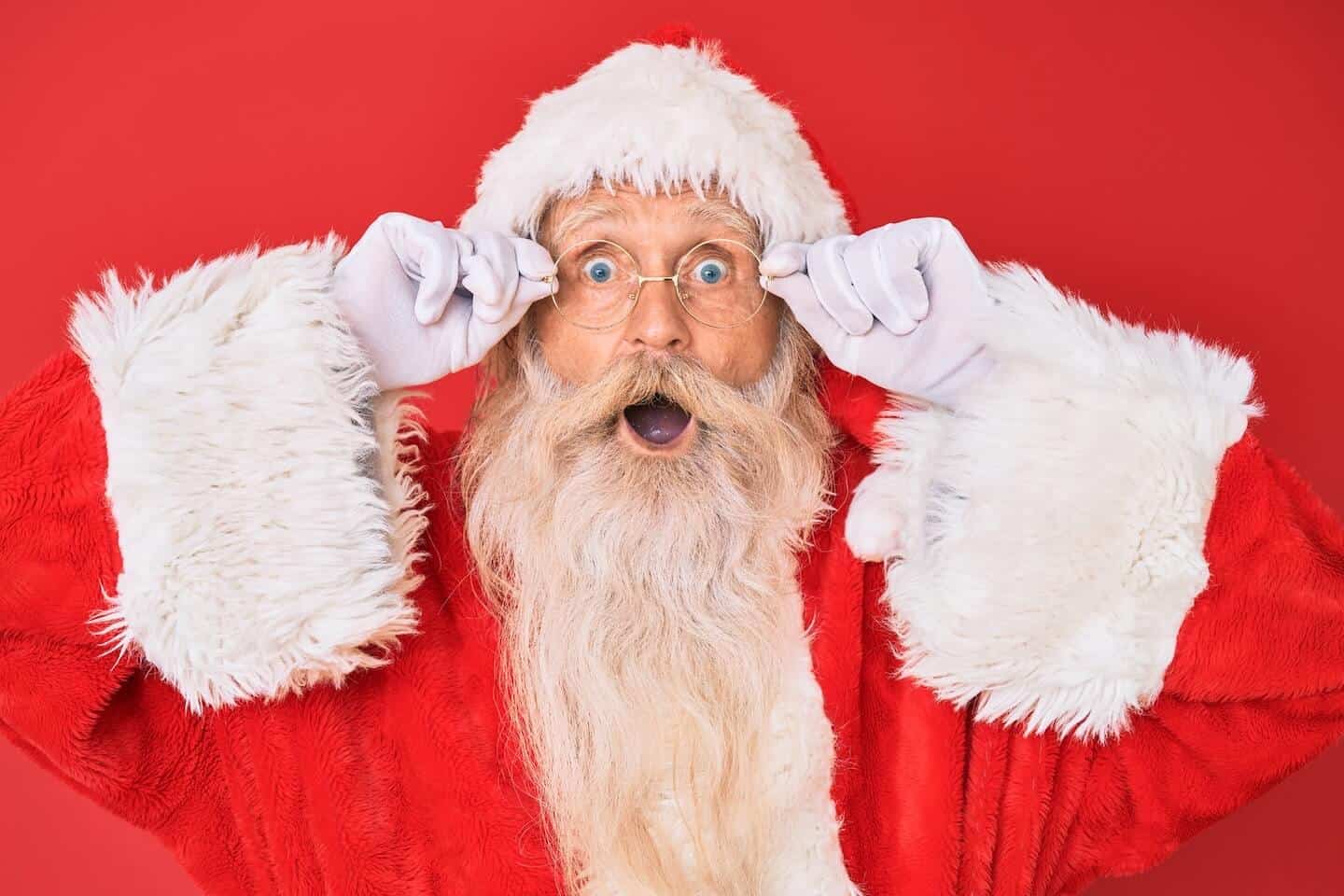 Santa Claus agarrando los lados de sus gafas con ambas manos y con la boca abierta mirando sorprendido.