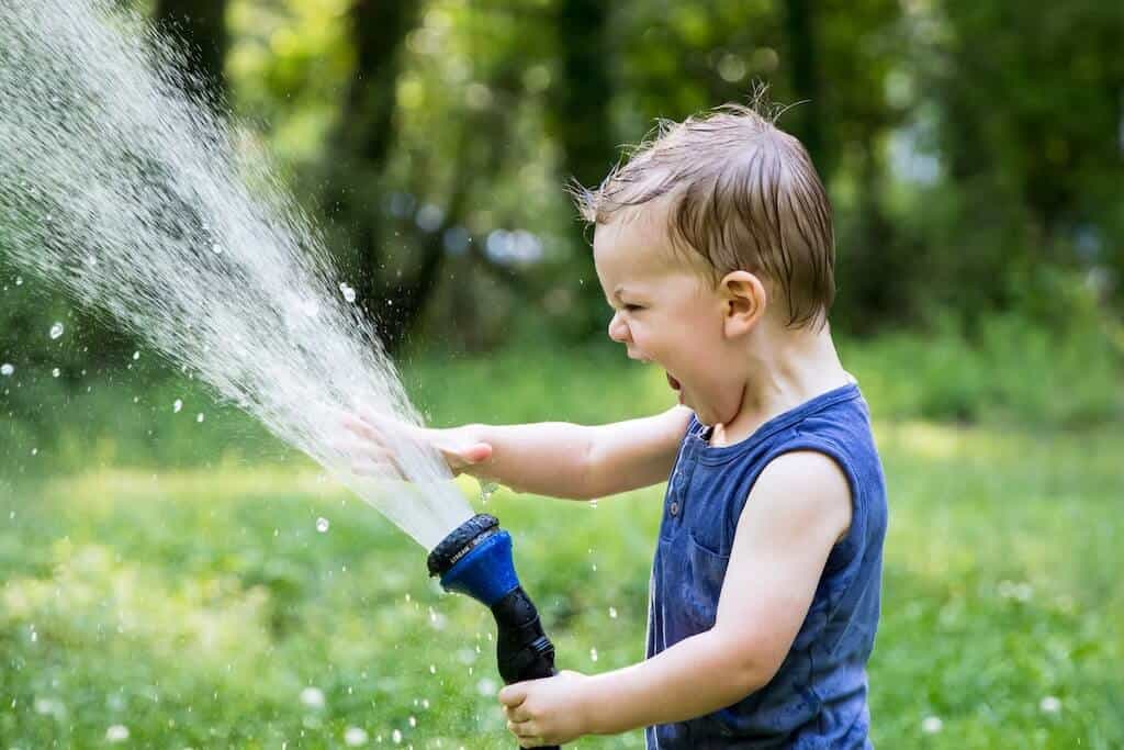 Niño rubio con una camiseta azul sosteniendo una manguera de jardín con agua saliendo de ella y poniendo su mano en la corriente de agua.
