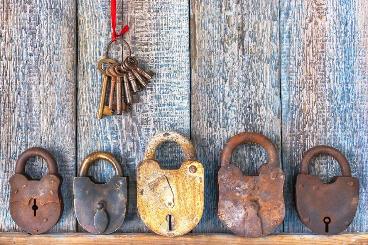 Cinco candados antiguos en varios colores apoyados contra una pared desgastada.  Sobre las cerraduras, un anillo de llaves cuelga de una cinta.